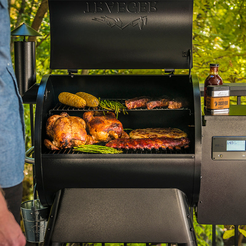 A black Traeger Pro575 outdoor pellet grill with the hood open, showing cooked chicken, steak, corn, asparagus, and ribs on the grill grate.