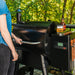 A black Traeger Pro 575 pellet smoker on an outdoor deck, a person is holding the front handle.