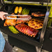A black Traeger Pro575 outdoor pellet grill with multiple foods cooking on the grates. A person's hand holding metal tongs is visible on the left.