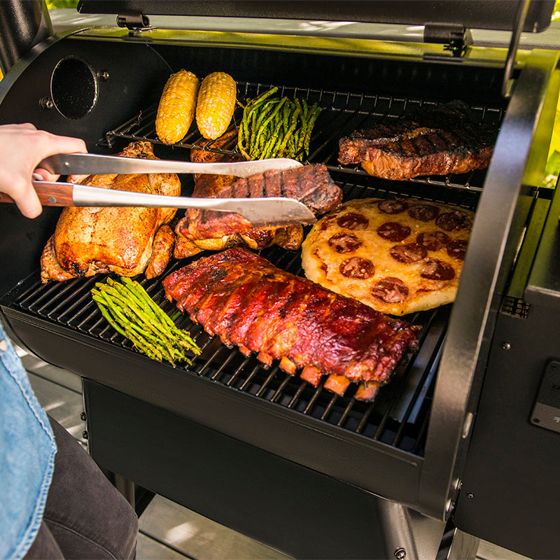 A black Traeger Pro575 outdoor pellet grill with multiple foods cooking on the grates. A person's hand holding metal tongs is visible on the left.